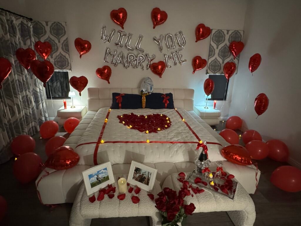 Proposal Airbnb room setup in San Jose with with "Will You Marry Me" balloons, heart-shaped fresh rose petals, curated champagne, framed pictures, and a dozen roses with glass vase
