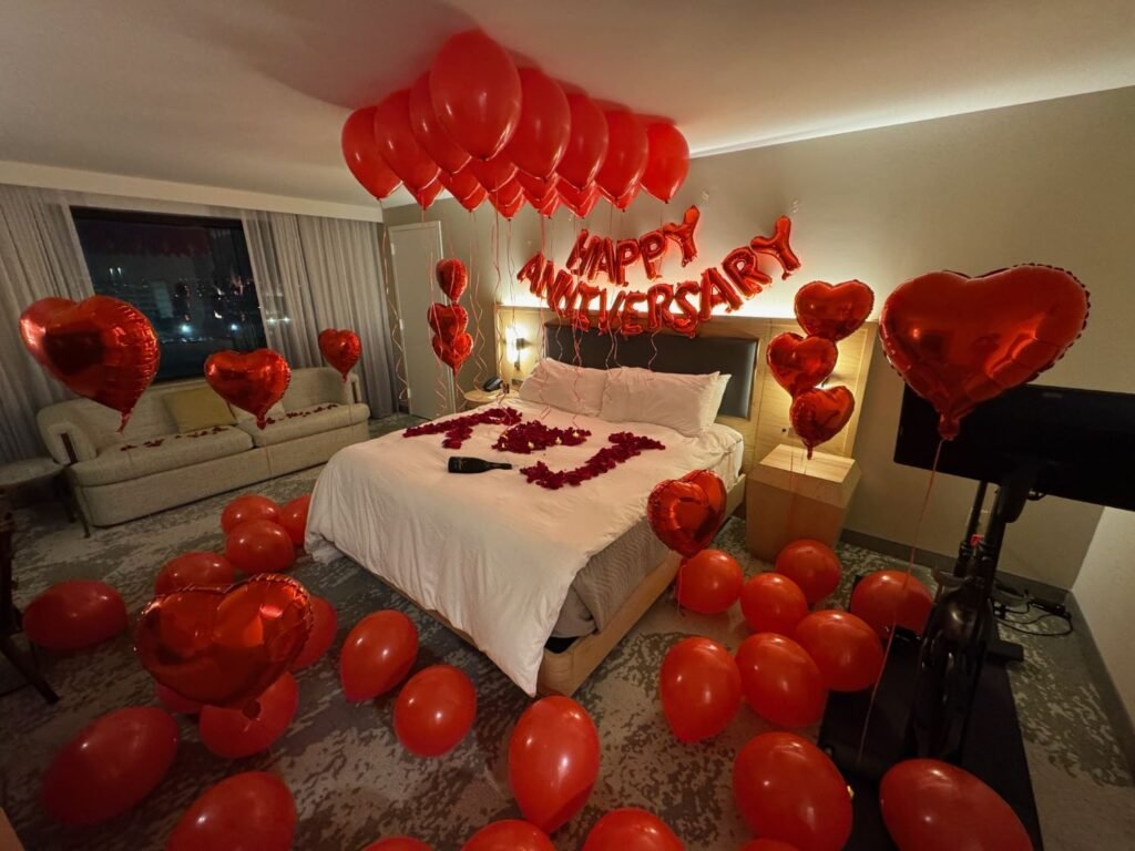 Romantic anniversary room decoration with candles, champagne, heart-shaped rose petals, balloon decor, curated wine, premium chocolate, and keepsake box