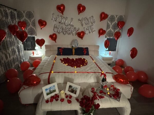 Proposal room setup in San Jose with with "Will You Marry Me" balloons, heart-shaped fresh rose petals, curated champagne, framed pictures, and a dozen roses with glass vase